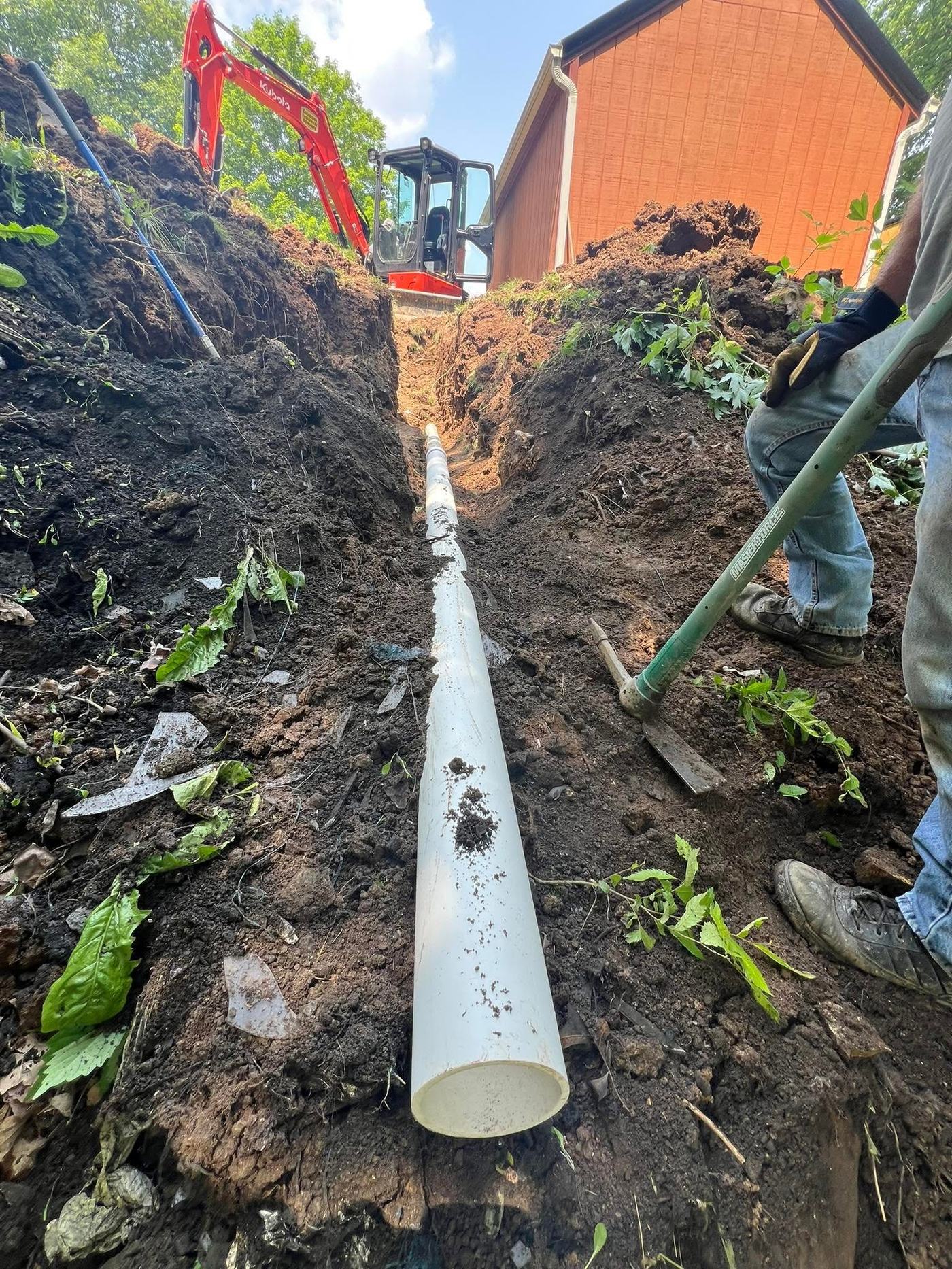 Waterline pipe laid in an open trench, with a Kubota mini excavator working at the far end near a barn